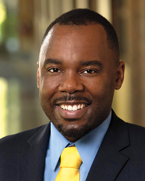 A young African American man in a suit coat wearing a blue dress shirt and yellow tie, smiling facing the camera.