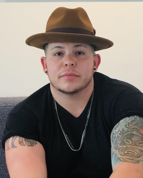 A head shot of Nic López Rodriguez wearing a black v-neck shirt with silver chains and a brown hat