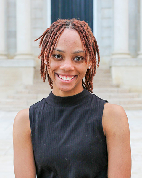 Headshot of Ani-ya Beasley, a young dark-skinned feminine person standing in front of a stone building with pillars