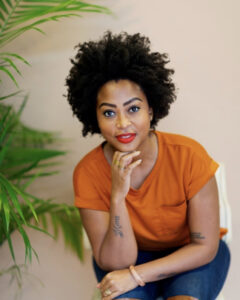 portrait of Stephanie D. Keene, a medium brown Black woman with an afro. she is wearing an orange shirt and resting her chin on her hand.