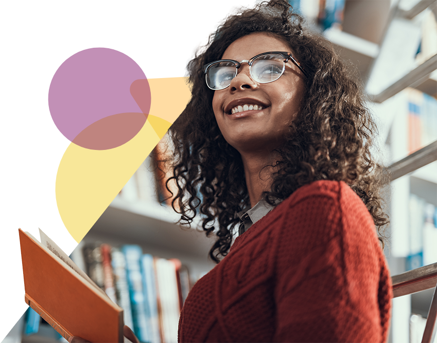 A dark skinned woman smiles while holding a book in front of a bookcase