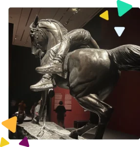A giant artistic sculpture of a horse that appears to be in stride and carrying a Black man who is positioned across the back of the horse with his legs hanging off of the back of the horse. The man is wearing a pair of Nike sneakers and jeans. In the background, there are burgundy-colored walls and people in the exhibit. A woman with black dreadlocked hair and glasses who is wearing black jeans, a black shirt, a blue jean jacket and brown dress shoes is looking at the statue. A woman with her back turned to the statue appears to be reading content in white font that is on a burgundy wall. A dark-colored bench is in the corner of the exhibit room.
