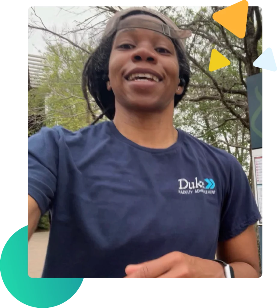 A Black woman with dreadlocks in a ponytail and a gray hat turned to the back is
wearing a dark-colored blue t-shirt that reads “Duke Faculty Advancement.” The woman, who is
wearing a Black Apple Watch and wedding ring is smiling and standing in front of a park sign
that reads Hermann Park and has a map. The Hermann Park sign with the accompanying map is
posted on three dark-colored green posts. There are trees in the background and in the distance,
vehicles can be seen in what appears to be a parking lot area.