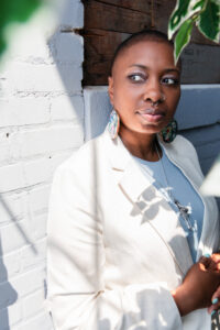 Dr. Dominique C. Hill, a black feminine person wearing all white leaning against a white brick wall with hanging greenery above zo