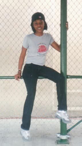 Kashema sitting outside of her high school on a green scaffolding wearing a backwards fitted cap over a chin length bob, smiling. She's wearing a gray t-shirt with red rose on the front with dark blue jeans and gray sneakers.