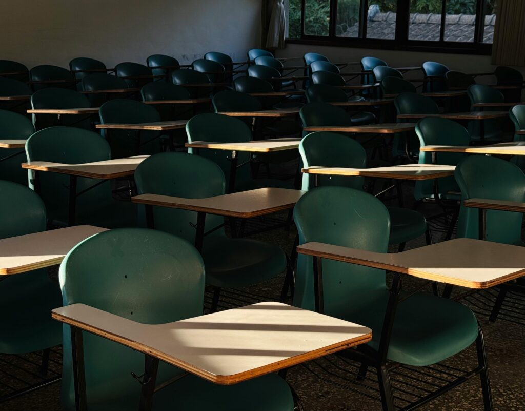 A dark classroom with several rows of empty tablet arm desks. The sunlight shines through the dark classroom, giving an ominous ambience.