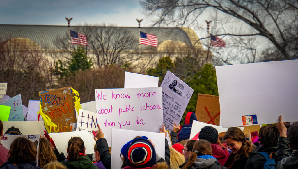 A group of people protests outside a government building with signs. One sign read, "We know more about public schools than you do."