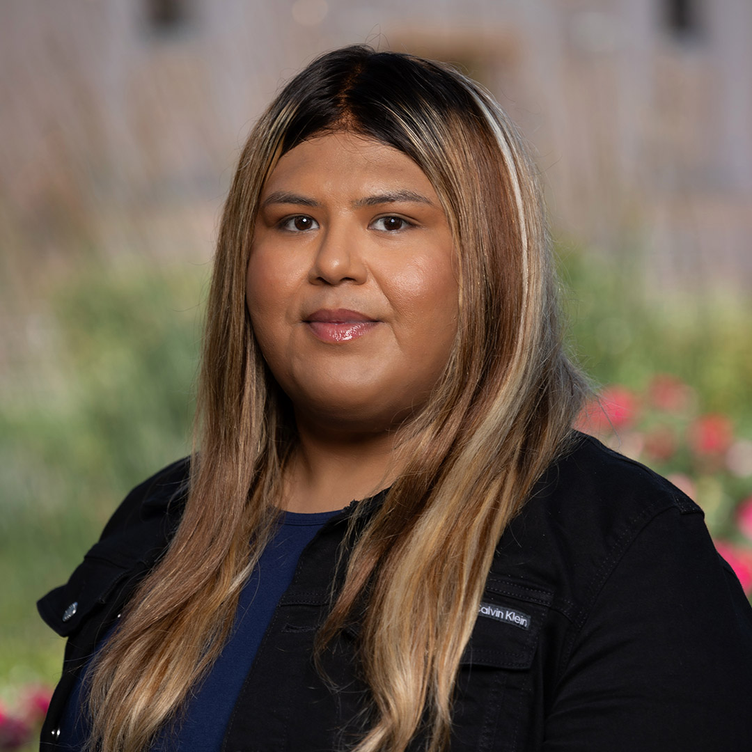 She is a brown woman with brown hair. She is wearing a blue top and a black jacket.