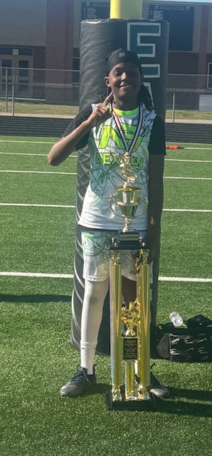 A, a pre-teen to teenaged black boy with a drug and sports apparel, stands holding up his fingers to indicate being number one while wearing a medal and posing with a trophy on a football field