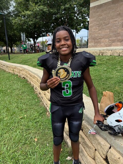 A, a pre-teen to teenaged black boy with braided hair and a football jersey, smiles while holding up a medal in front of a stone wall