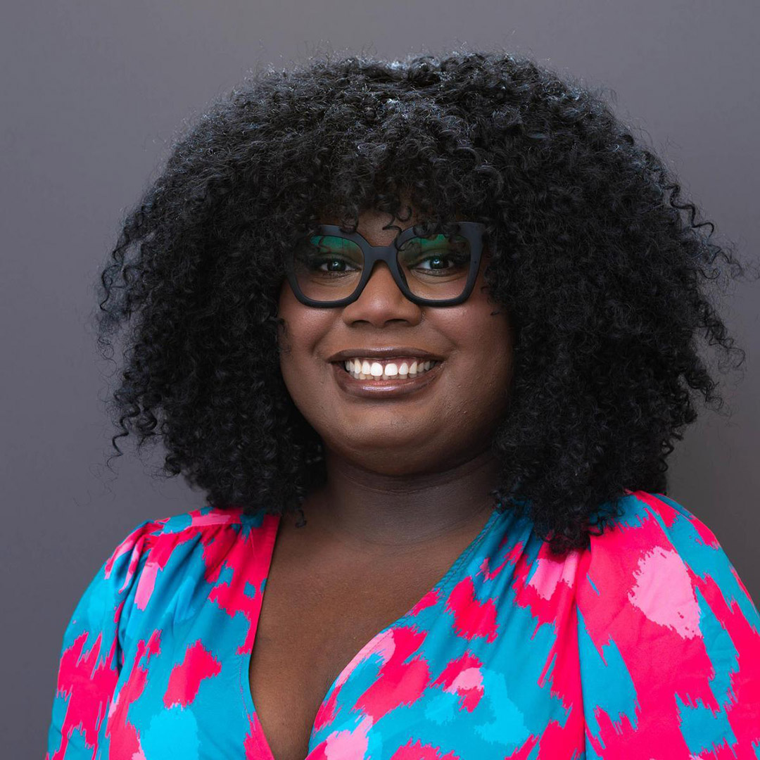 She is a Black woman with kinky black hair. She is wearing a pink-and-blue dress with black glasses and standing in front of a gray background.