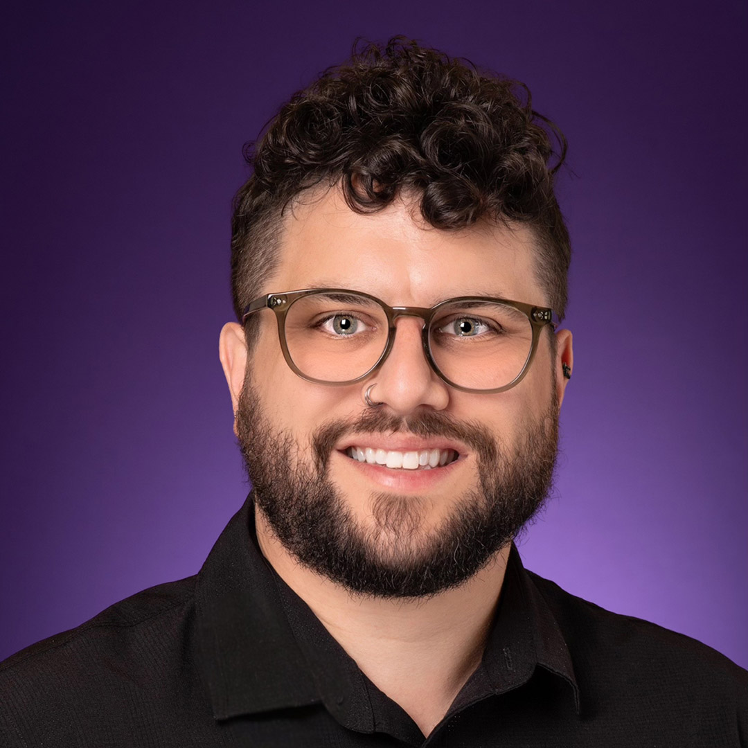 Dr. Gavin P. Johnson (he/him). He is a white man with curly brown hair and a short beard. He is wearing glasses and a black shirt. There is a purple background.