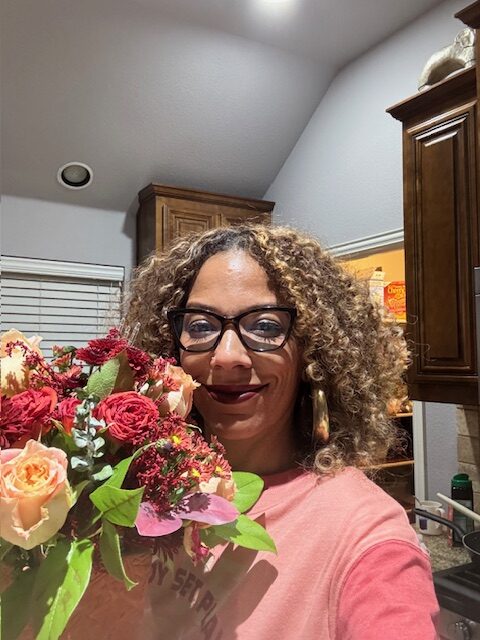 Kelly Franklin, a black woman with curly blonde hair, glasses and a pink shirt, holds a bouquet of flowers.